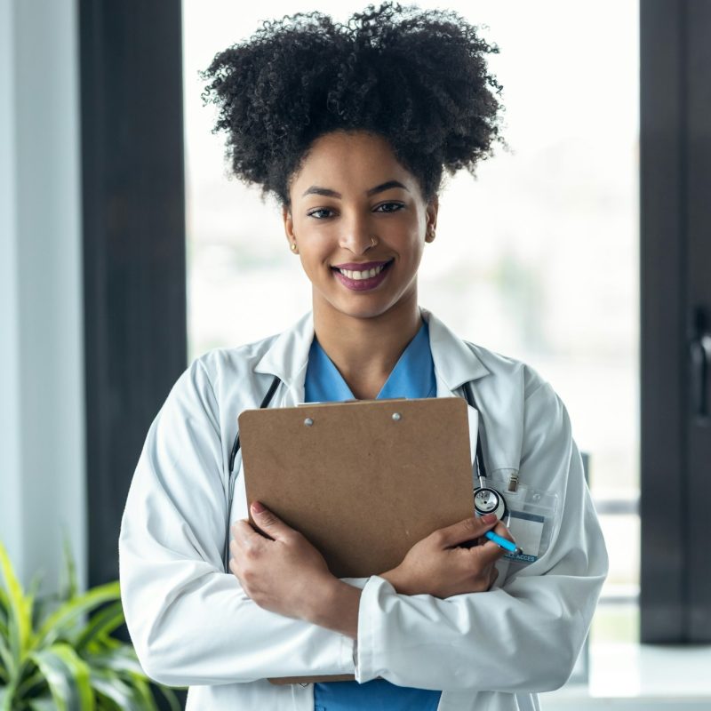 Afro female doctor holding clipboard while looking at camera standing in the consultation.